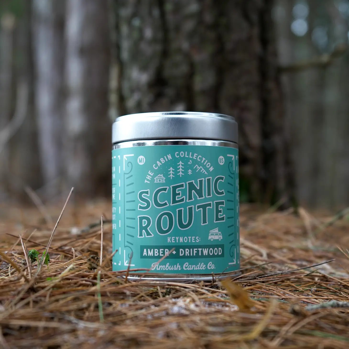 A tinned candle on a forest floor in turquoise blue and called ‘scenic route’ featuring the scents amber, driftwood.