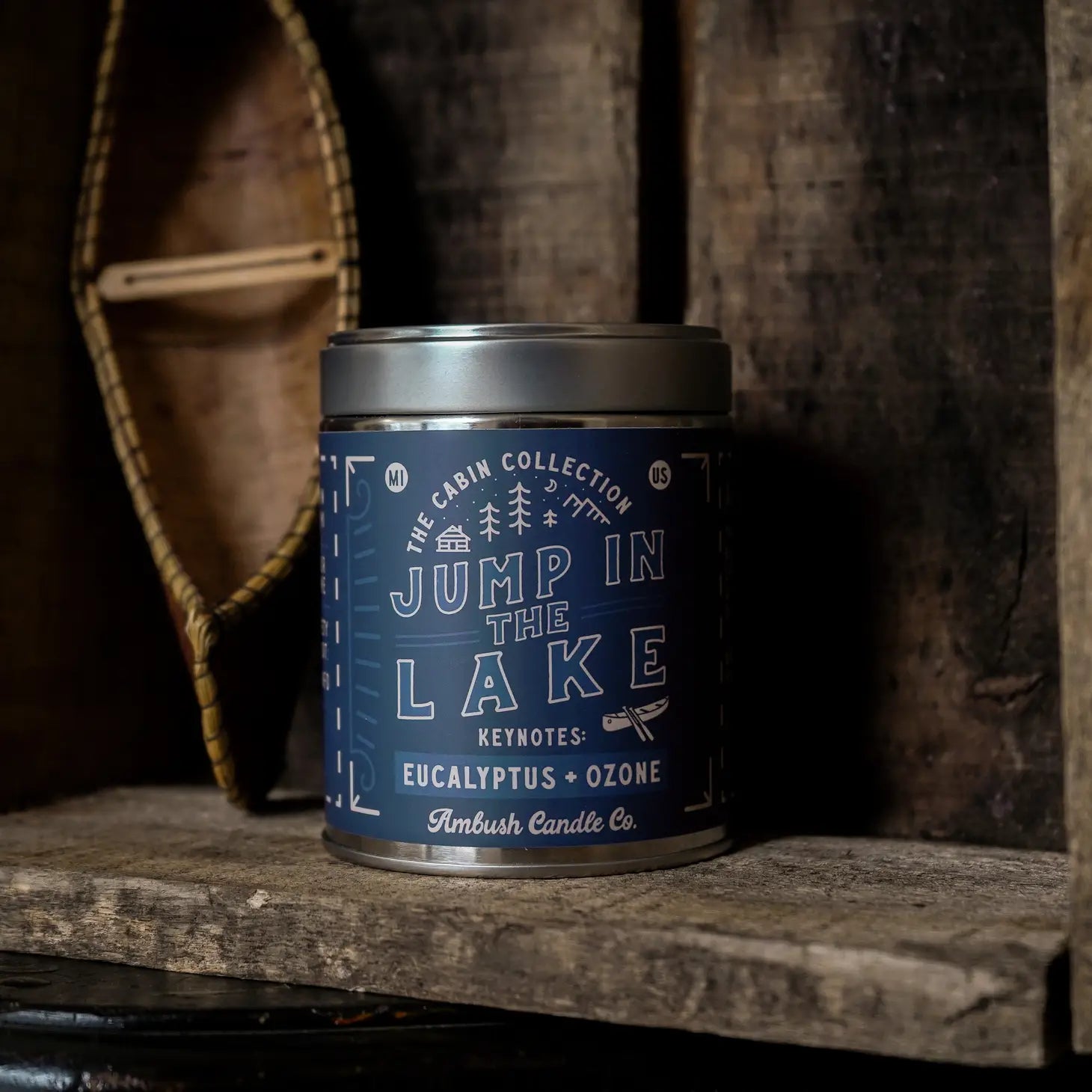 A 14oz blue scented candle tin labeled "Ambush Candle Co Jump In The Lake" with notes of eucalyptus and ozone, made from vegan soy wax, sits on a rustic wooden shelf.