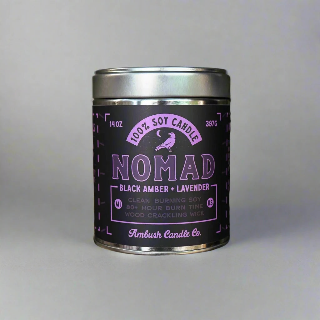 A tin candle labeled "Ambush Candle Co 14OZ Nomad Black Amber & Lavender," crafted from clean-burning soy wax, is nestled between two tree trunks in a serene forest setting.