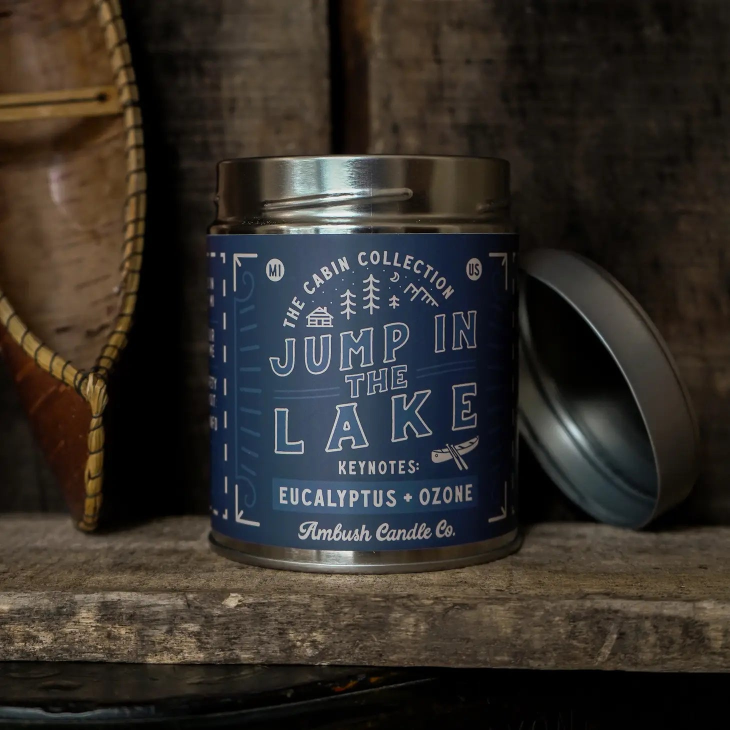 A 14oz blue scented candle tin labeled "Ambush Candle Co Jump In The Lake" with notes of eucalyptus and ozone, made from vegan soy wax, sits on a rustic wooden shelf.