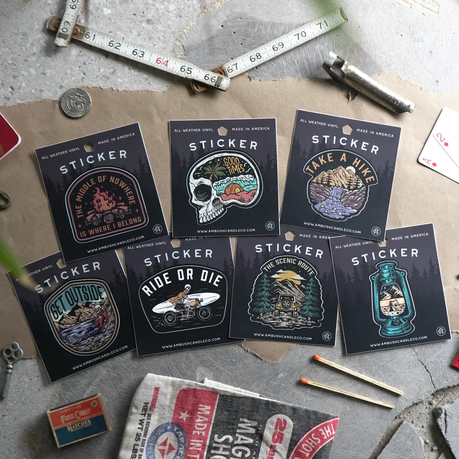 The Ambush Candle Co Ride or Die Sticker, featuring an assortment of weatherproof vinyl designs with outdoor and adventure motifs, is displayed amidst measuring tools, coins, and playing cards on a rustic surface. Made in the USA and dishwasher safe for lasting durability.