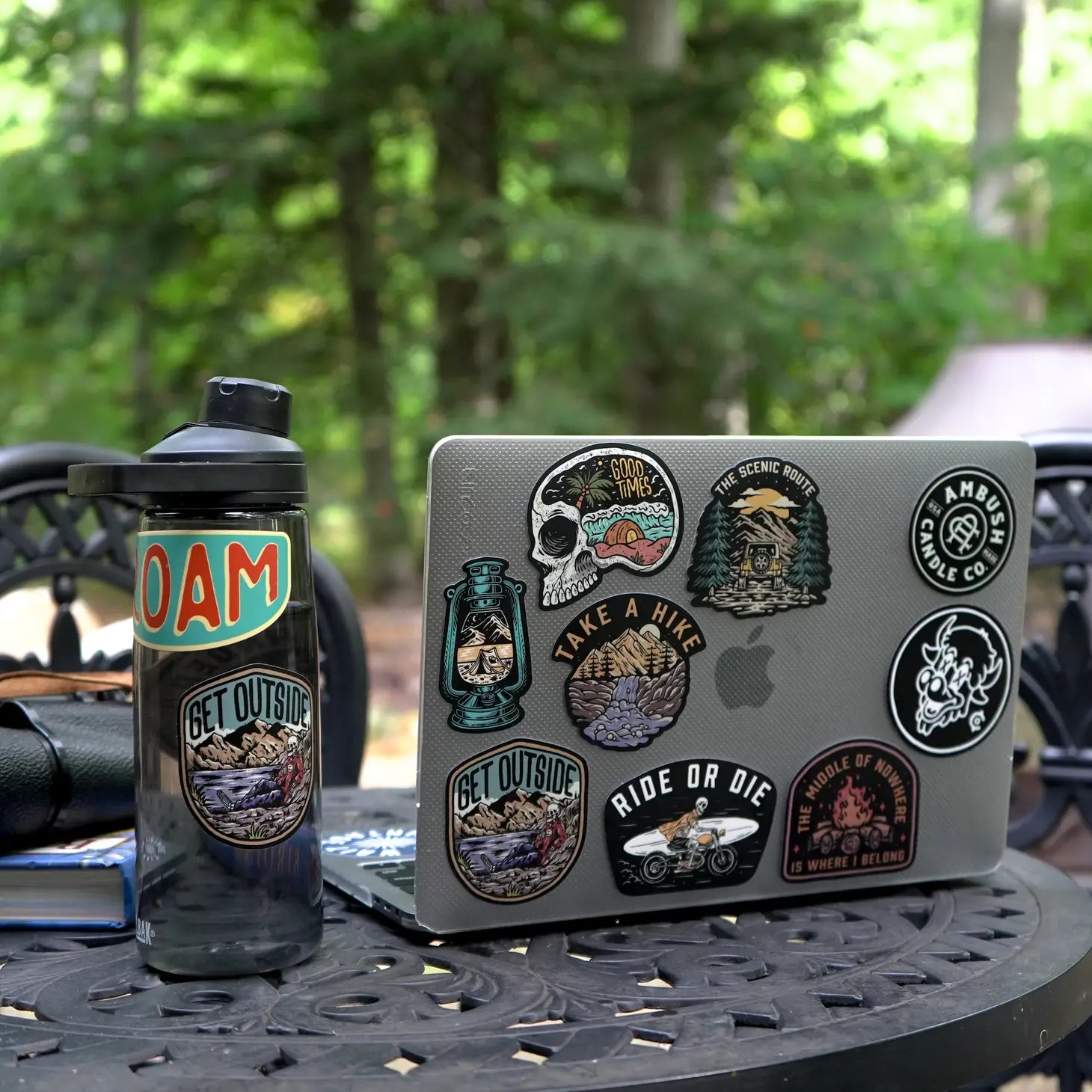 A laptop featuring an assortment of outdoor-themed decals, including the Ambush Candle Co Ride or Die sticker, and a water bottle adorned with weatherproof vinyl stickers, all made in the USA, rest on a table outdoors in a forested area.