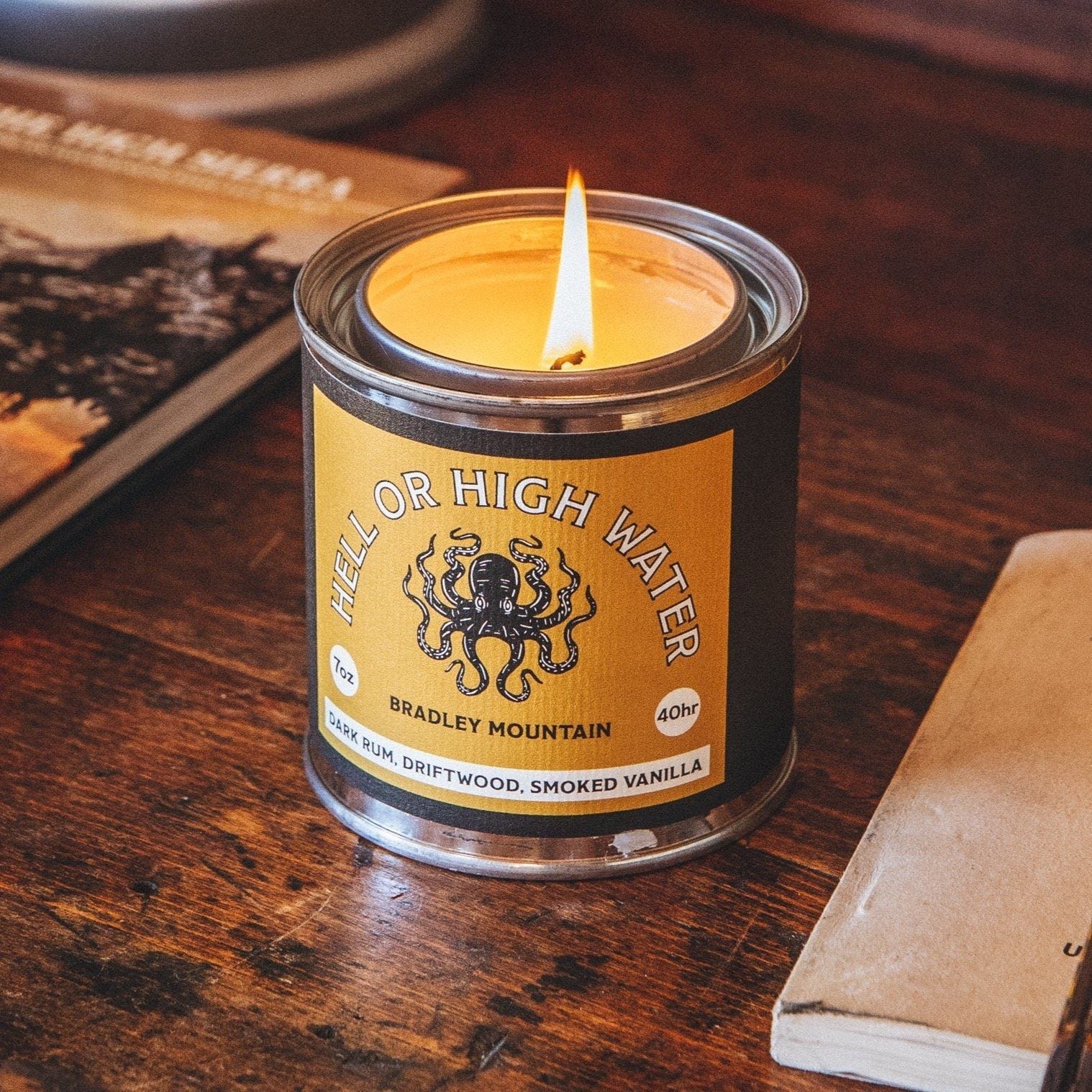 A candle tin labeled "Bradley Mountain Hell Or High Water" with an octopus graphic. The Bradley Mountain Hell Or High Water Dark Rum, Driftwood & Smoked Vanilla Candle features the enticing scent of dark rum, driftwood, and smoked vanilla. Burn time: 40 hours.