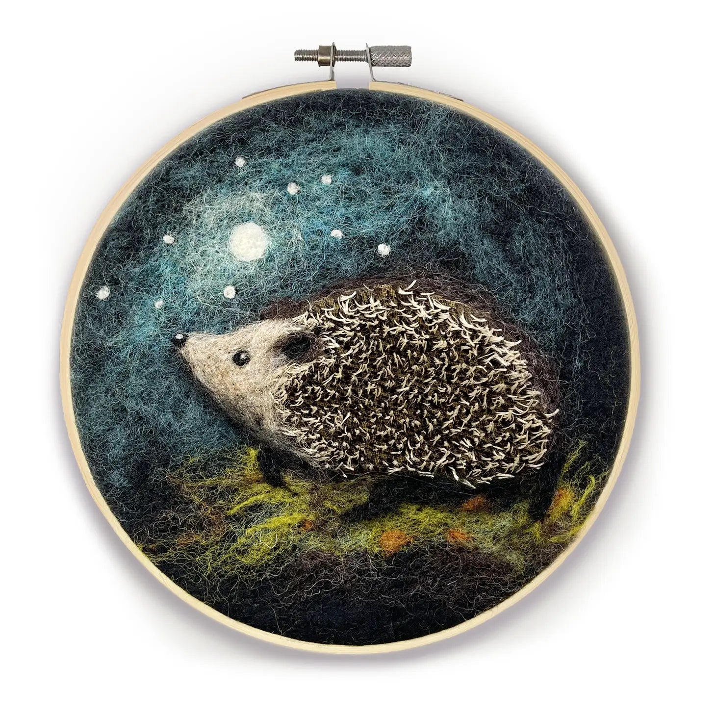 A felt artwork of a hedgehog under a moonlit night sky, displayed in an embroidery hoop. The Crafty Kit Company's "Hedgehog In A Hoop" Needle Felting Craft Kit is perfect for beginners looking to explore the art of needle felting.