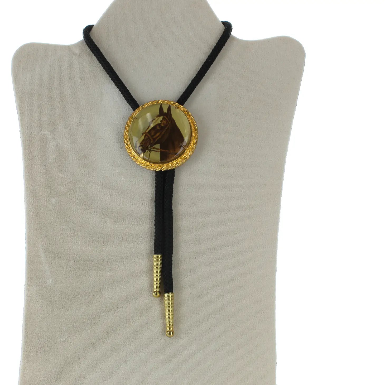 The Forager Provisions Horse Portrait Bolo Tie is hand-made and displayed on a white stand, featuring a large, gold and brown decorative slide. It also includes black braided cords with gold tips and an adjustable cord for a perfect fit.