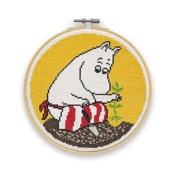 The Crafty Kit Company Moominmamma Gardening Cross Stitch features a cross-stitched artwork of a white Moomin character gardening, set in a circular embroidery hoop with a yellow background. Made using high-quality embroidery thread, this piece is perfect for fans of Moominmamma and comes as part of a complete cross stitch kit.