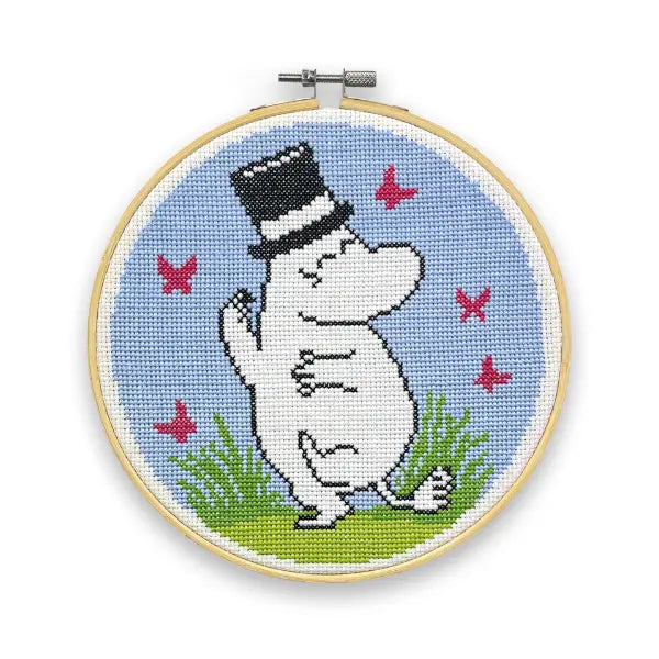 The Crafty Kit Company Moominpapa Dancing Cross Stitch features an embroidery of a white Moomin, specifically Moominpappa, adorned with a black top-hat. The design showcases a blue background, pink butterflies, and green grass, all beautifully framed in a hoop. It is perfect for those following the beginners' guide to cross stitch.