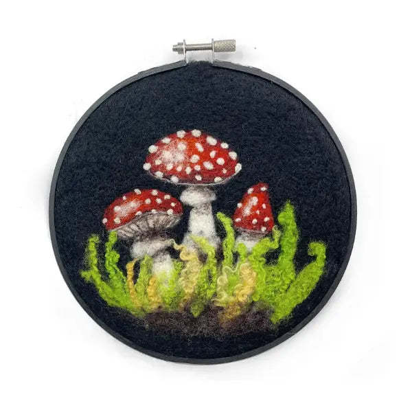 The Crafty Kit Company Mushrooms In A Hoop features an intricate embroidery of red and white mushrooms surrounded by green foliage, all beautifully framed in a circular black embroidery hoop.